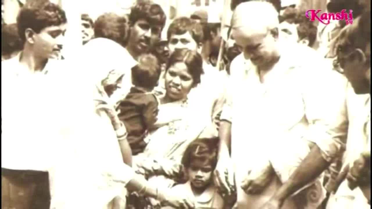 A crowd surrounds a man in a white shirt, and a woman in a sari holds a child. The Kanshi TV logo is in the corner.
