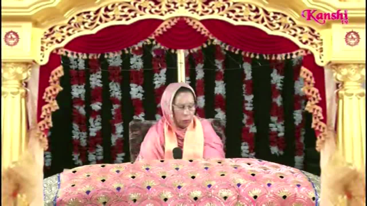 A woman in a pink head covering speaks into a microphone, seated behind a table decorated with a peacock feather pattern. The Kanshi TV logo sits in the upper right corner, and the backdrop is a decorative stage setting.
