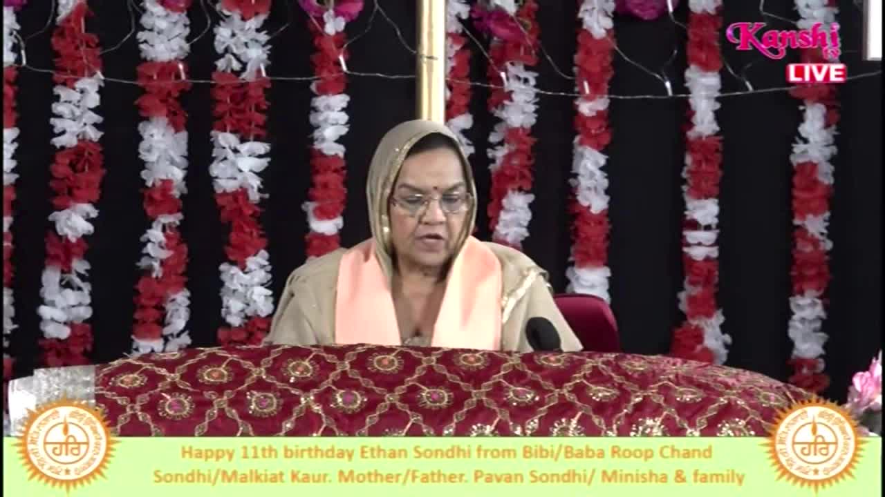 A woman in a head covering speaks into a microphone, her words broadcast live on Kanshi TV. Behind her, red and white garlands hang against a dark backdrop, and a message celebrates a birthday in the United Kingdom.

