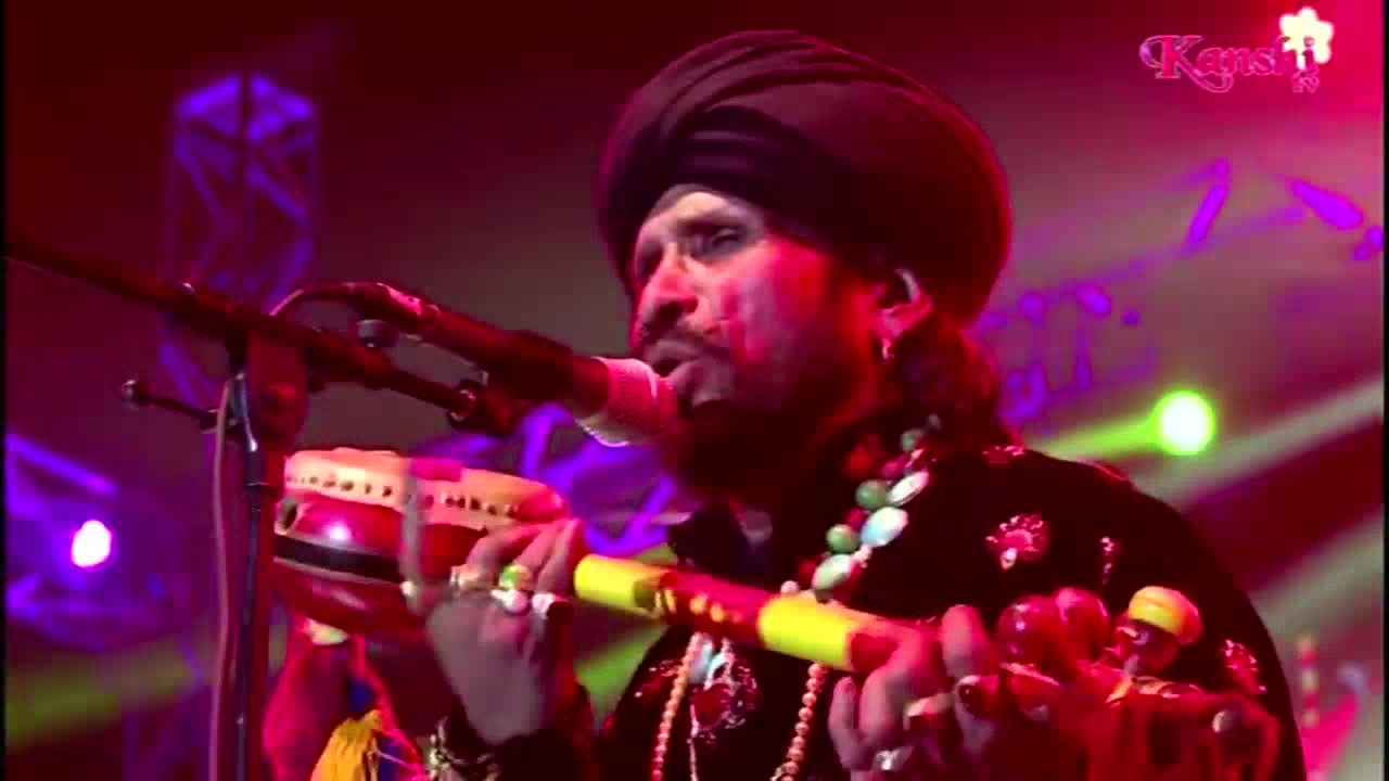 A man in a turban sings into a microphone, his hands moving over a colorful wind instrument and a small drum. The Kanshi TV logo is visible in the corner, and the stage glows with purple and pink lights.

