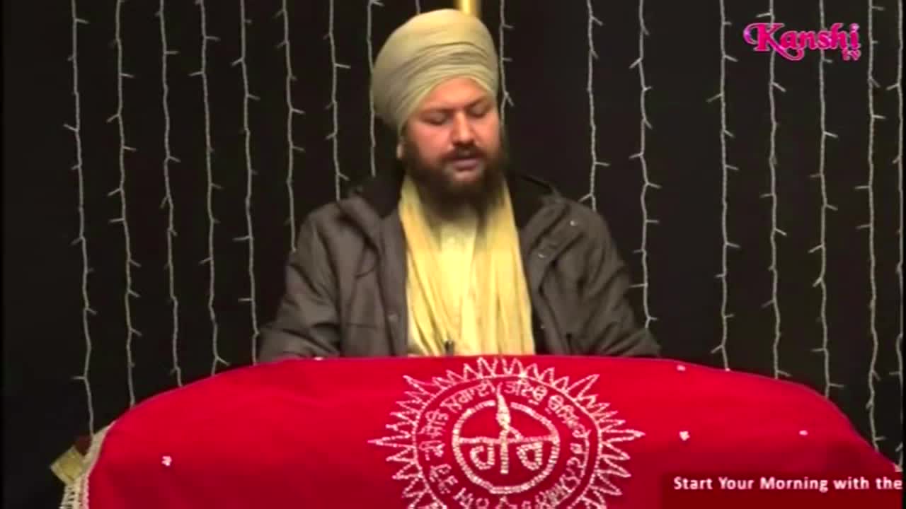A man in a turban and coat sits behind a red cloth, speaking. The Kanshi TV logo is visible in the corner, and strings of lights hang behind him.
