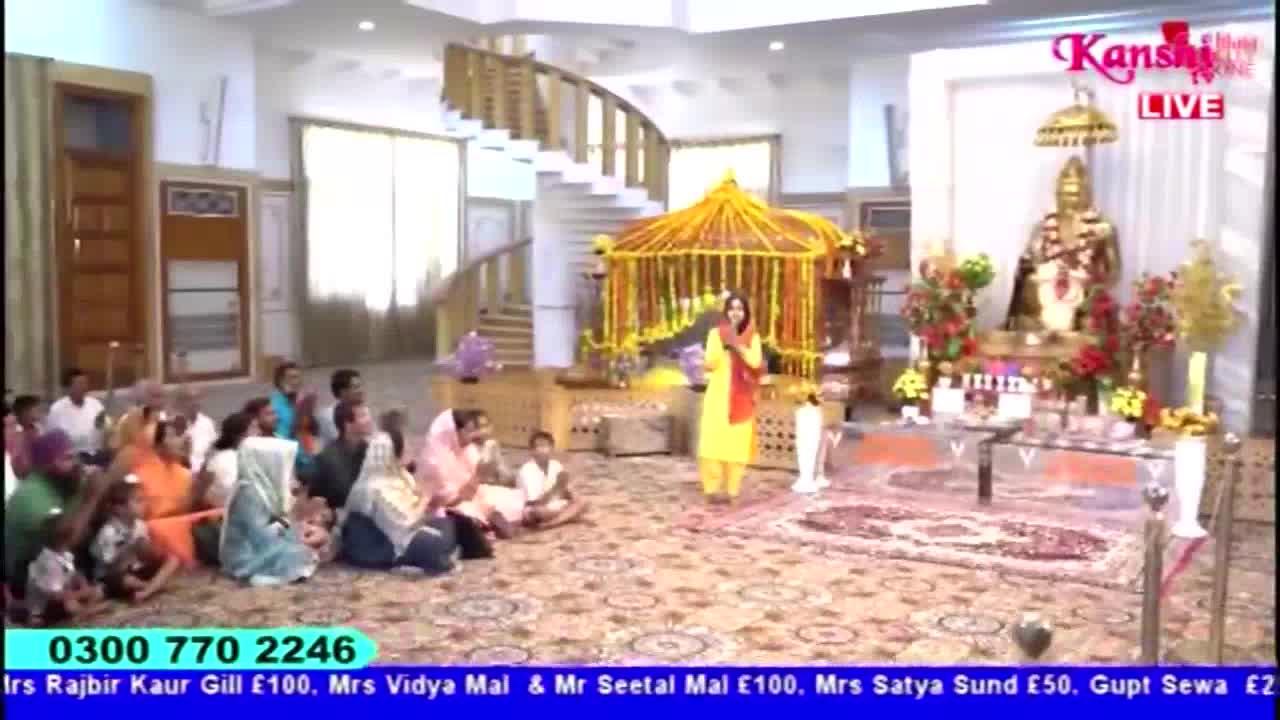 A woman in a yellow suit and red scarf speaks in front of a small crowd, with a gilded structure and an ornate statue behind her. The Kanshi TV logo is visible, and a phone number scrolls at the bottom of the screen.

