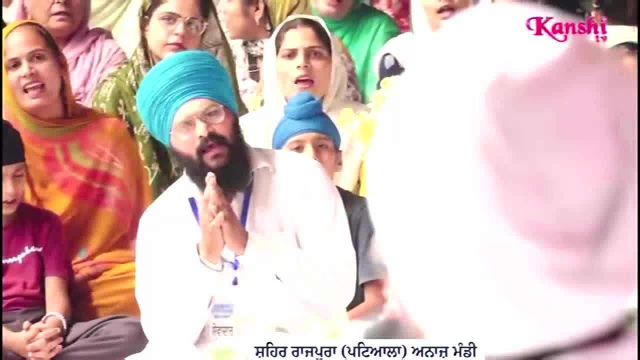 A man in a turquoise turban has his hands clasped in prayer, surrounded by others singing. The Kanshi TV logo is visible in the corner.

