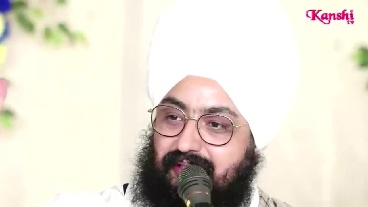 A man with a full beard and glasses speaks into a microphone, likely on Kanshi TV in the United Kingdom. He's wearing a white turban and his eyes are focused, as if he's addressing a crowd.
