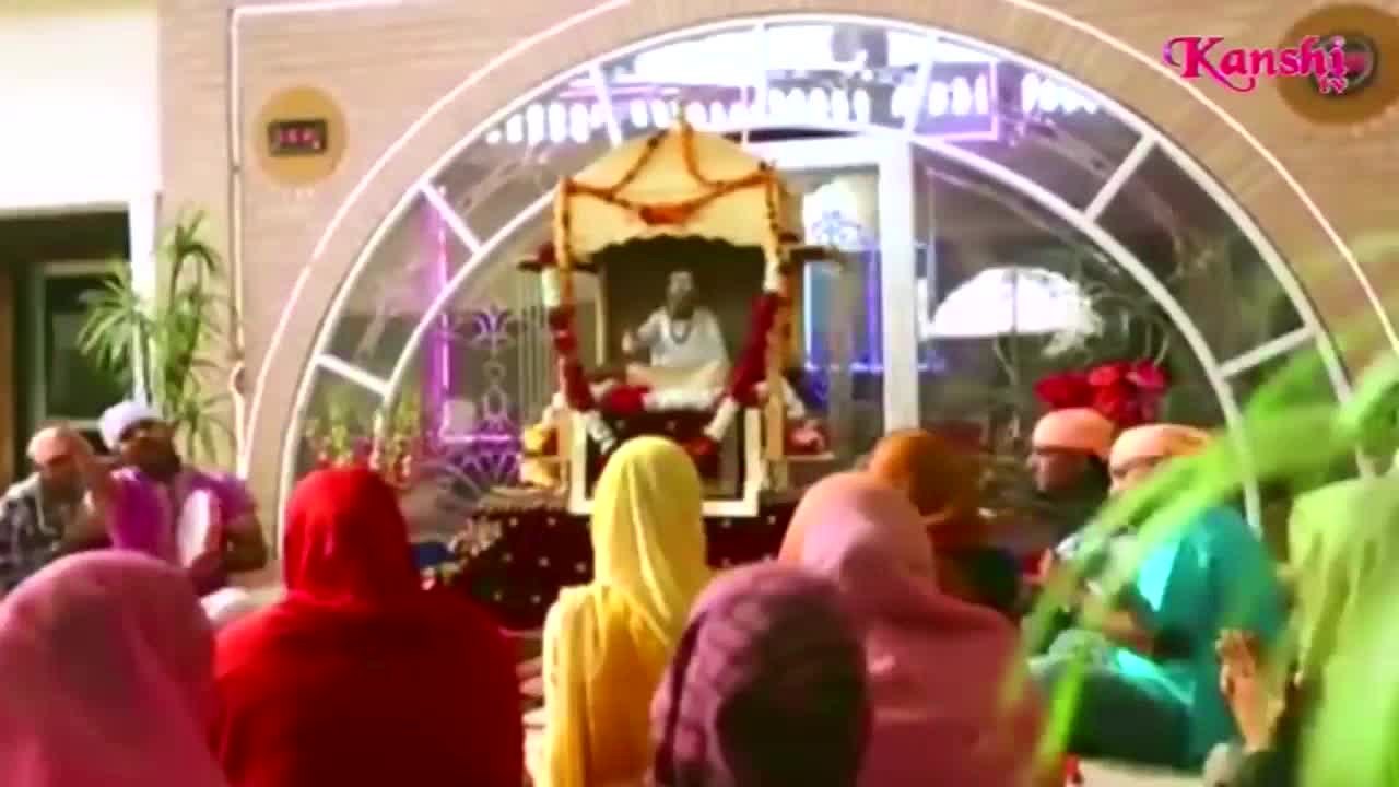 A robed figure sits elevated in an ornate, glass-enclosed structure, likely for a Kanshi TV broadcast. The audience, mostly women in colorful head coverings, faces the speaker.
