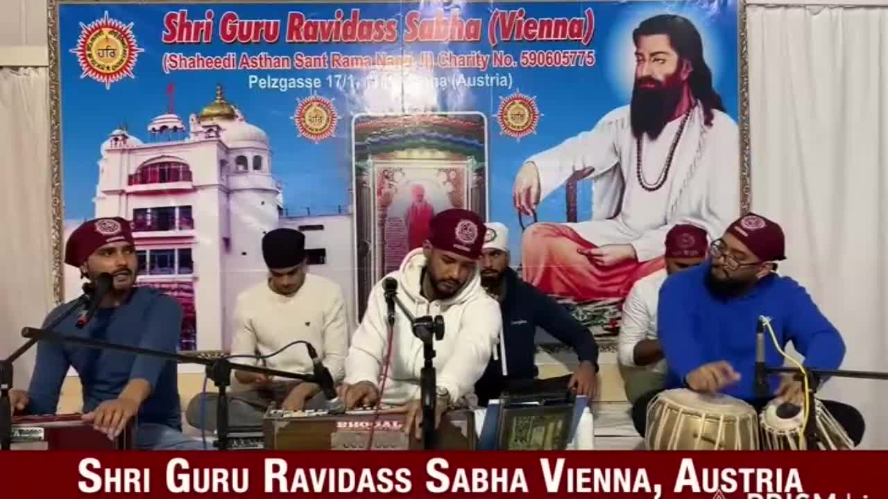 A group of men in maroon caps and casual clothes are seated, performing music with instruments like a harmonium and tabla. Behind them, a large banner displays text and an image of a religious figure.

