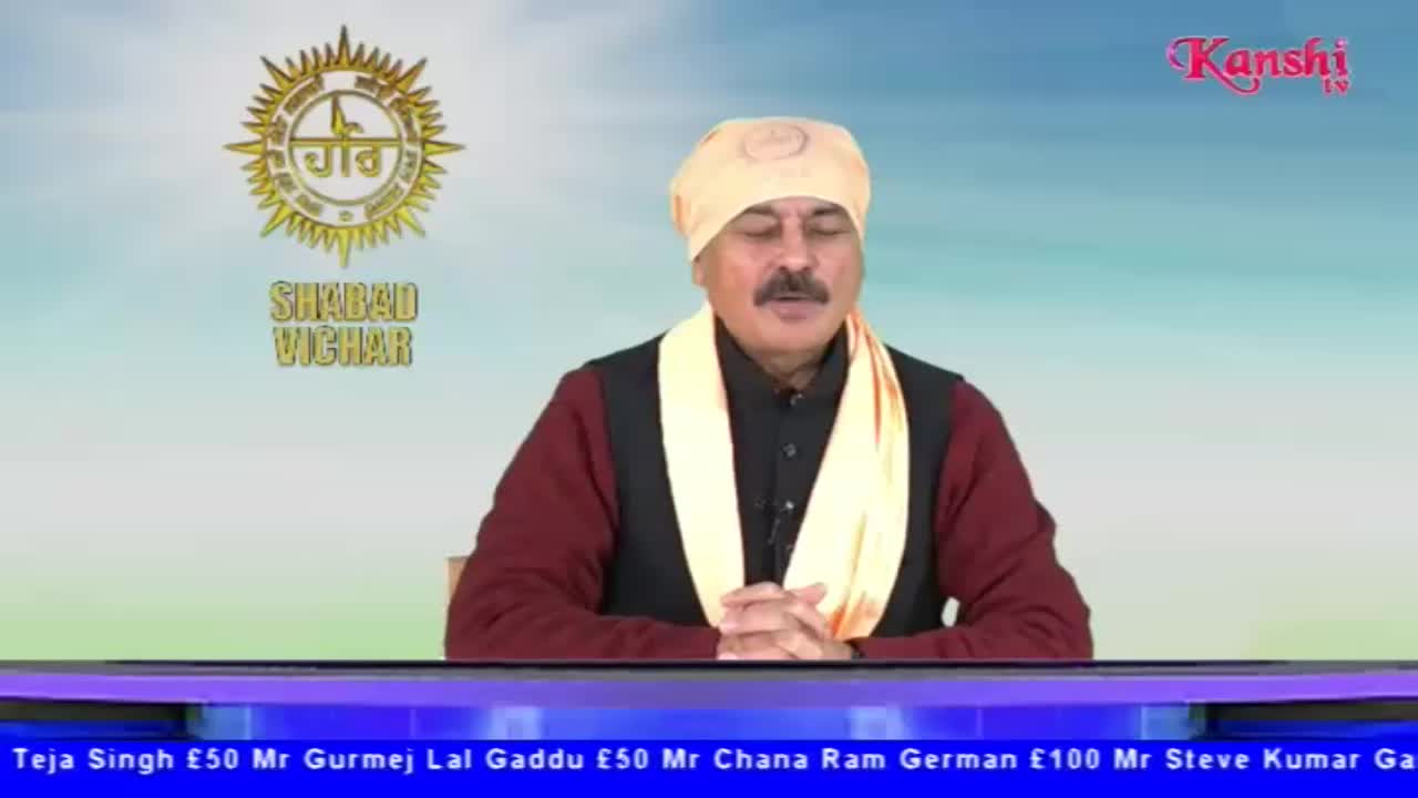 A man in a turban and shawl sits at a desk, speaking on Kanshi TV. The backdrop features a logo and a soft blue gradient.
