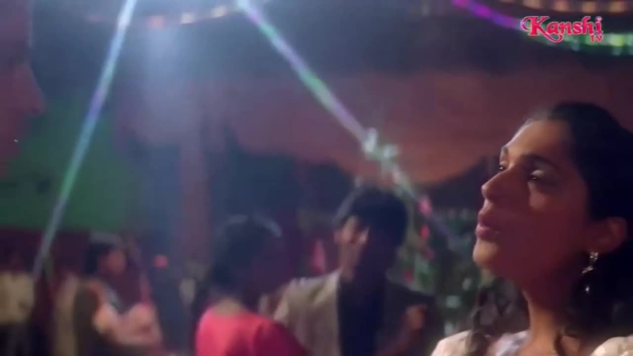 A woman in a white top looks off to the side, illuminated by a spotlight. Behind her, blurry figures dance under the Kanshi TV logo.
A woman in a white top looks off to the side, illuminated by a spotlight. Behind her, blurry figures dance under the Kanshi TV logo.