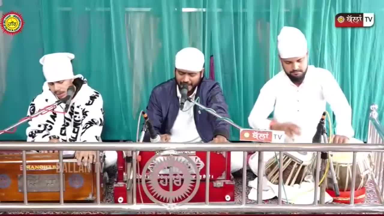 Three men, all wearing white turbans, sit behind instruments, performing. The center man sings into a microphone, while the others play a harmonium and tabla drums, respectively.
Three men, all wearing white turbans, sit behind instruments, performing. The center man sings into a microphone, while the others play a harmonium and tabla drums, respectively.