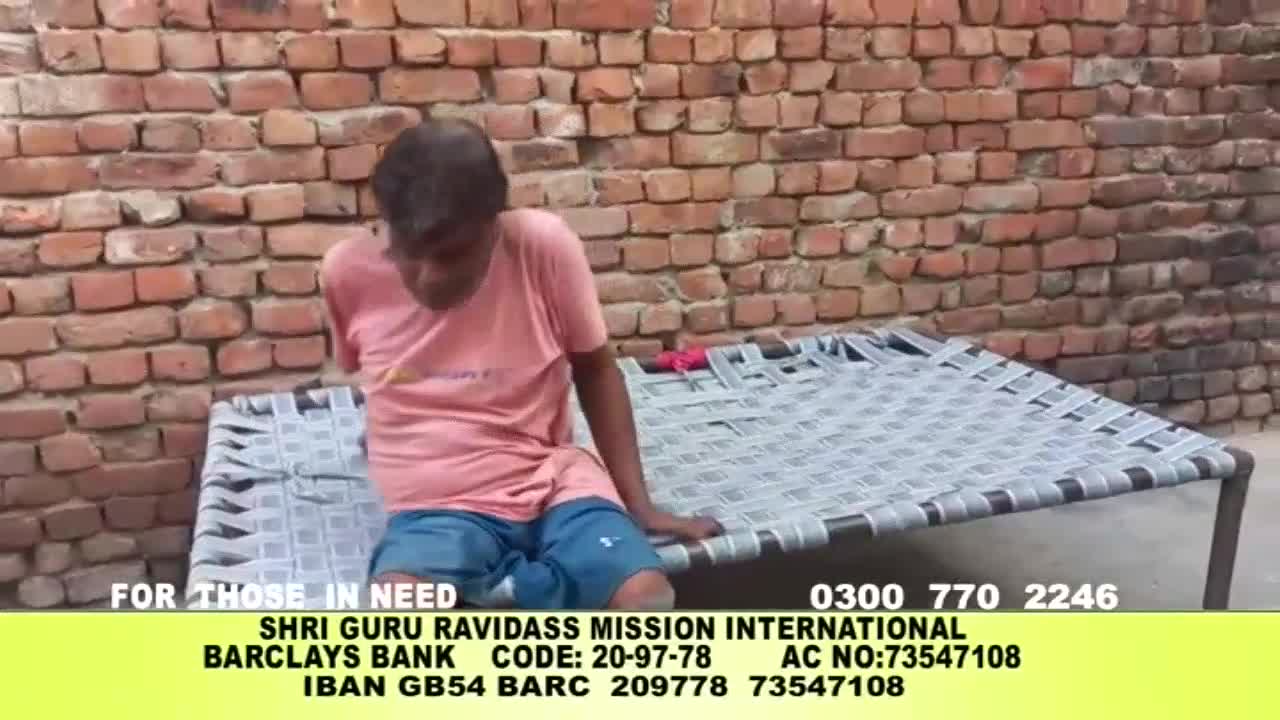 A man with no arms sits on a woven bed frame, looking down. The Kanshi TV screen shows a plea for donations, with contact information for a charity in the United Kingdom.
