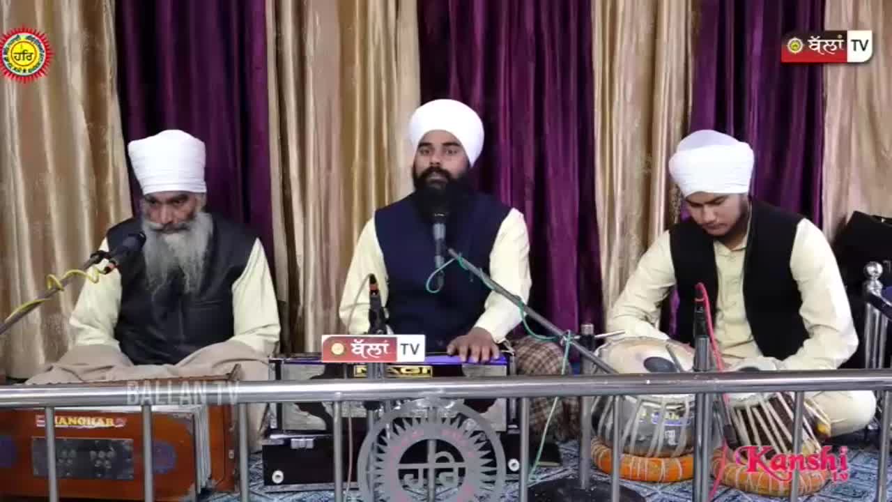Three men in turbans and vests are seated behind musical instruments, with microphones in front of them. The center man is speaking, while the man on the right plays the tabla.
