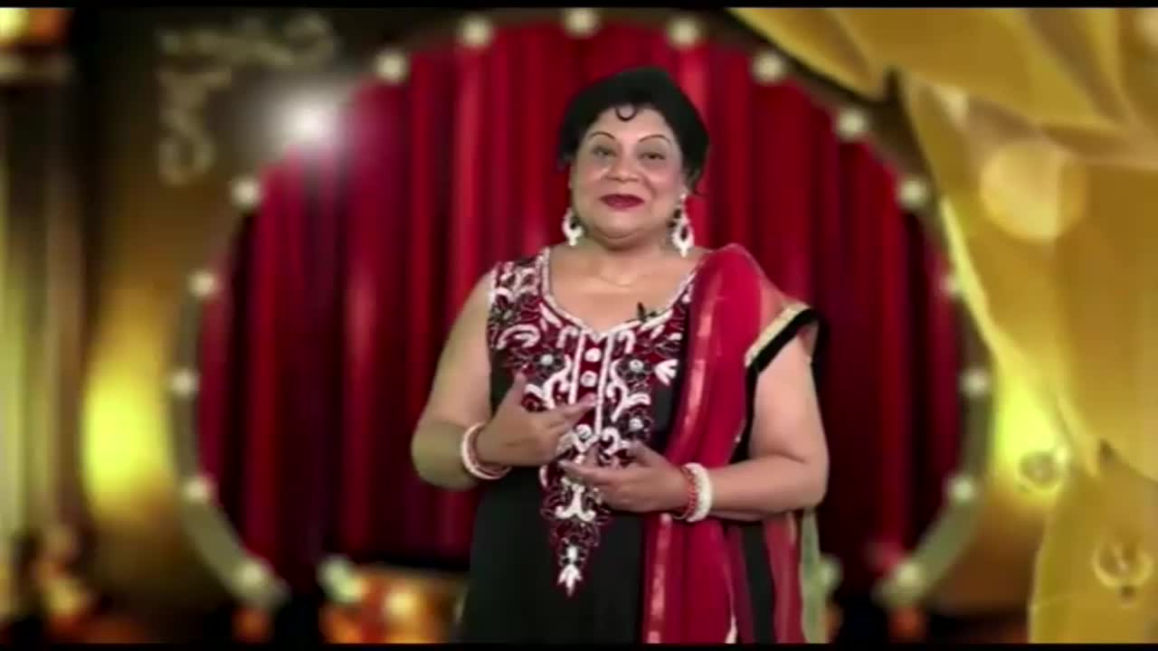 A woman in a black and red outfit stands center stage, gesturing with her hands. Behind her, a red curtain and gold accents frame the scene, likely for a Kanshi TV show in the United Kingdom.
