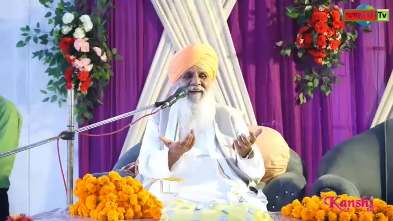 A man with a long white beard and orange turban speaks into a microphone, gesturing with his hands. Kanshi TV's logo is visible on the stage, where marigold garlands are arranged. 
