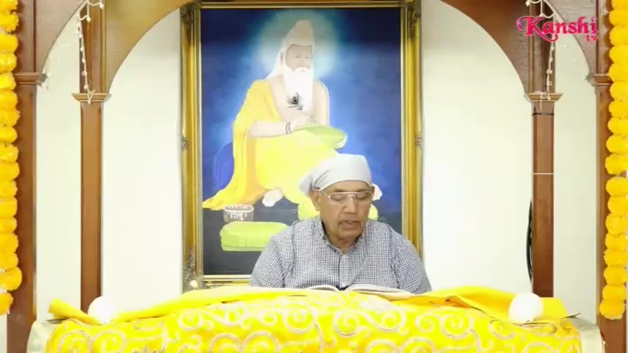 A man in glasses and a white head covering reads from a book, positioned in front of a painting. The Kanshi TV logo is visible in the corner, and the scene is framed with yellow floral garlands.
