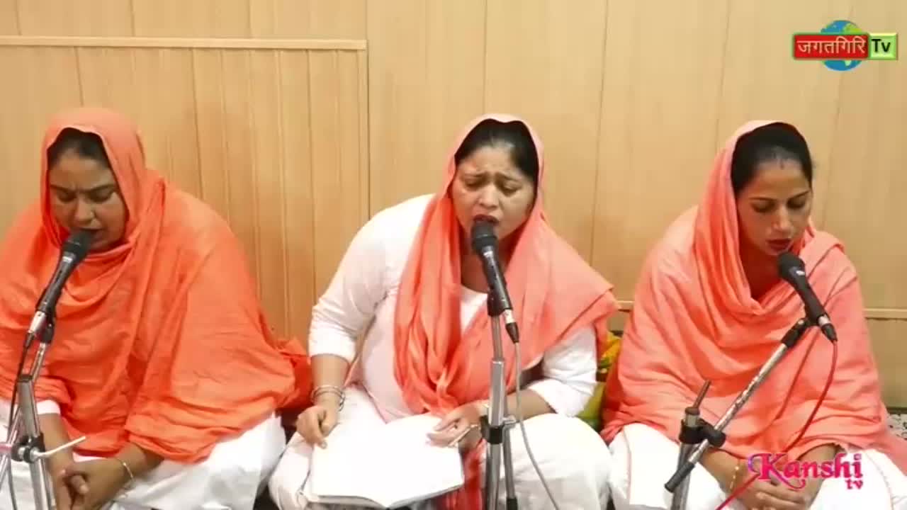 Three women, each wearing an orange head covering and white clothing, sing into microphones. The Kanshi TV logo is visible in the lower right corner.
Three women, each wearing an orange head covering and white clothing, sing into microphones. The Kanshi TV logo is visible in the lower right corner.
