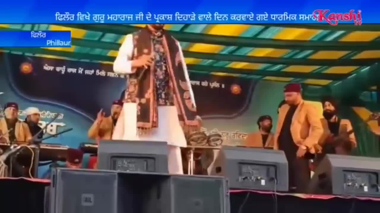 A man in a white kurta and patterned shawl stands at a microphone on a stage. To his right, another man in a tan jacket walks past, and musicians play instruments behind them.