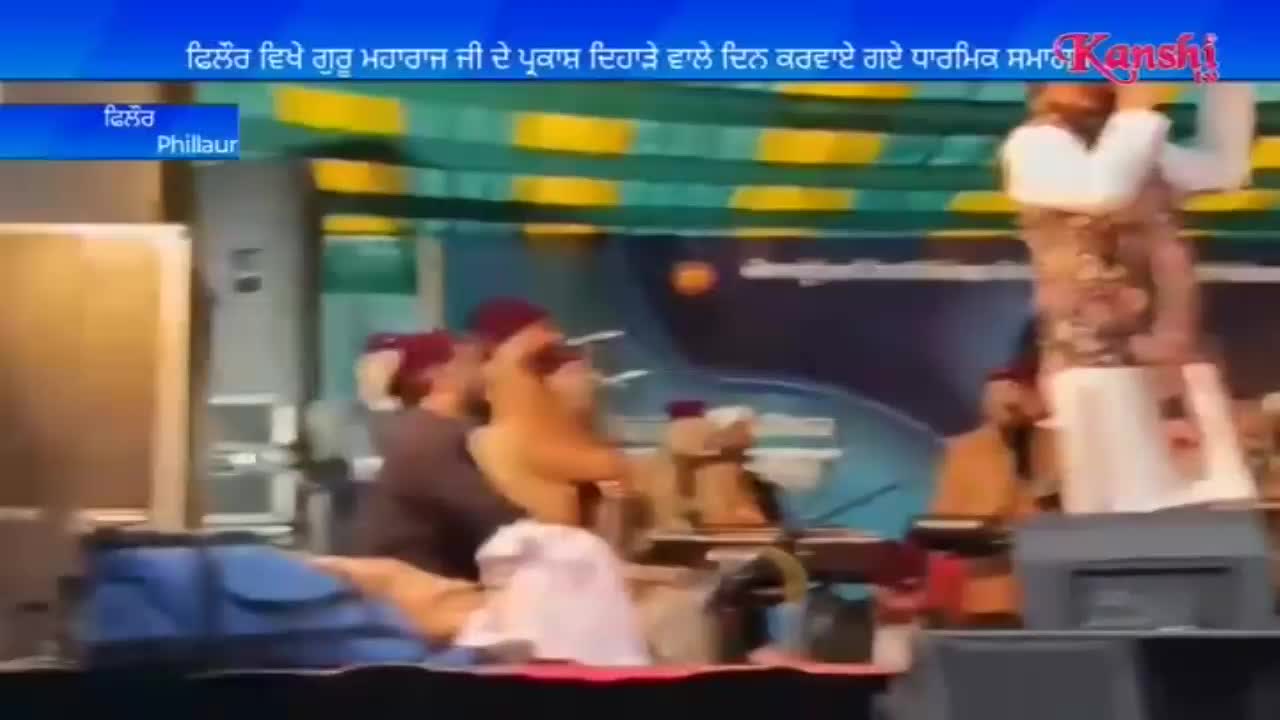 A man in a white tunic and patterned vest stands on a stage, holding a microphone aloft. Several other men, some in turbans, are seated behind him, playing instruments.