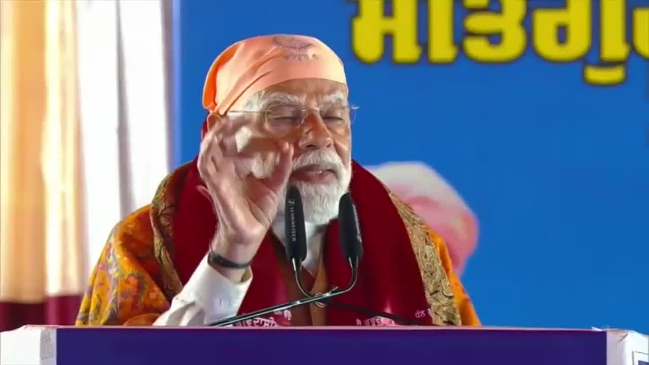 The speaker gestures with his right hand, his eyes closed, as he addresses the audience. A bright orange turban and a patterned shawl frame his face, with microphones positioned before him.