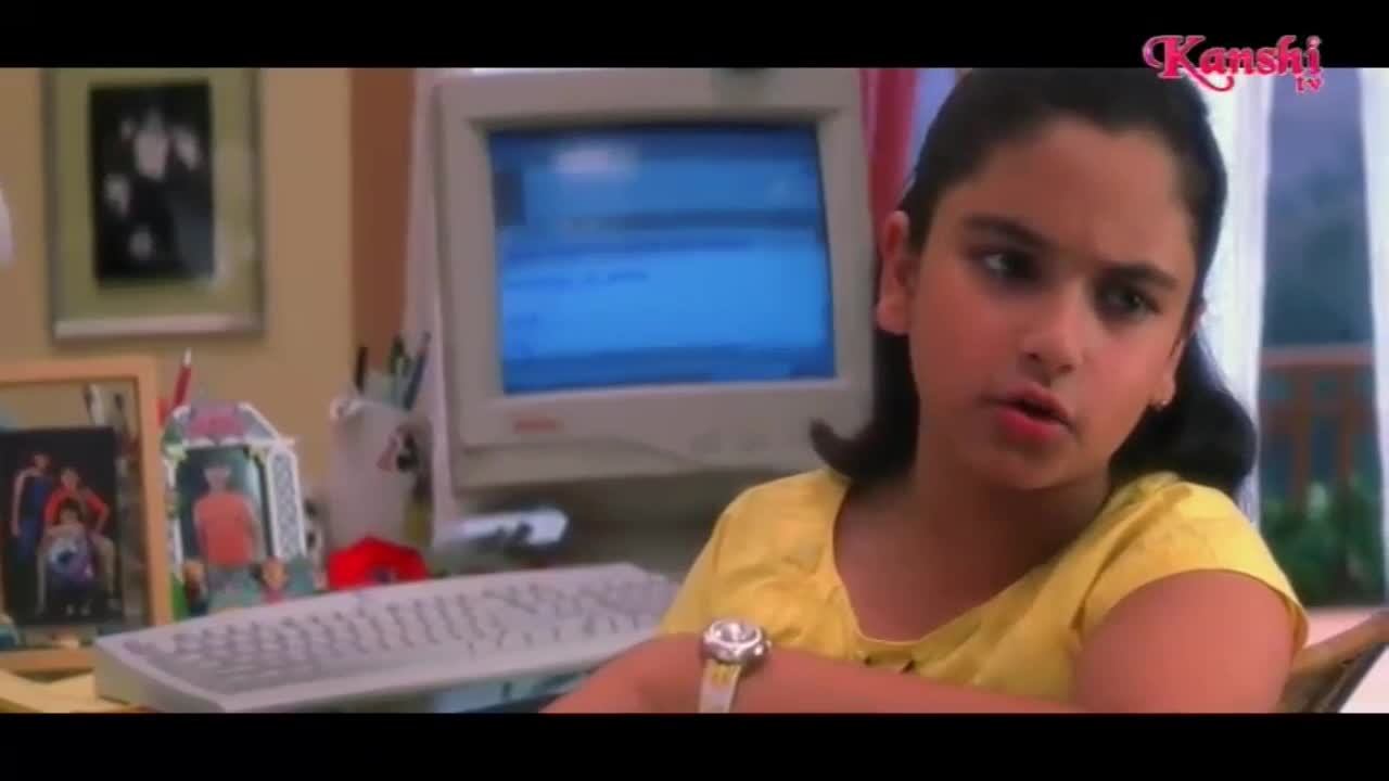 A young girl in a yellow top sits at a desk, her gaze fixed on something off-camera. The blue screen of an old computer monitor glows behind her, reflecting the room's light.