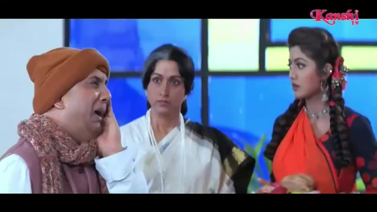 A man in a brown cap and scarf is speaking with his hand to his cheek, looking distressed. Behind him, two women stand with neutral expressions, one in a white sari and the other in a vibrant orange and blue outfit. The Kanshi TV logo appears in the top right corner.