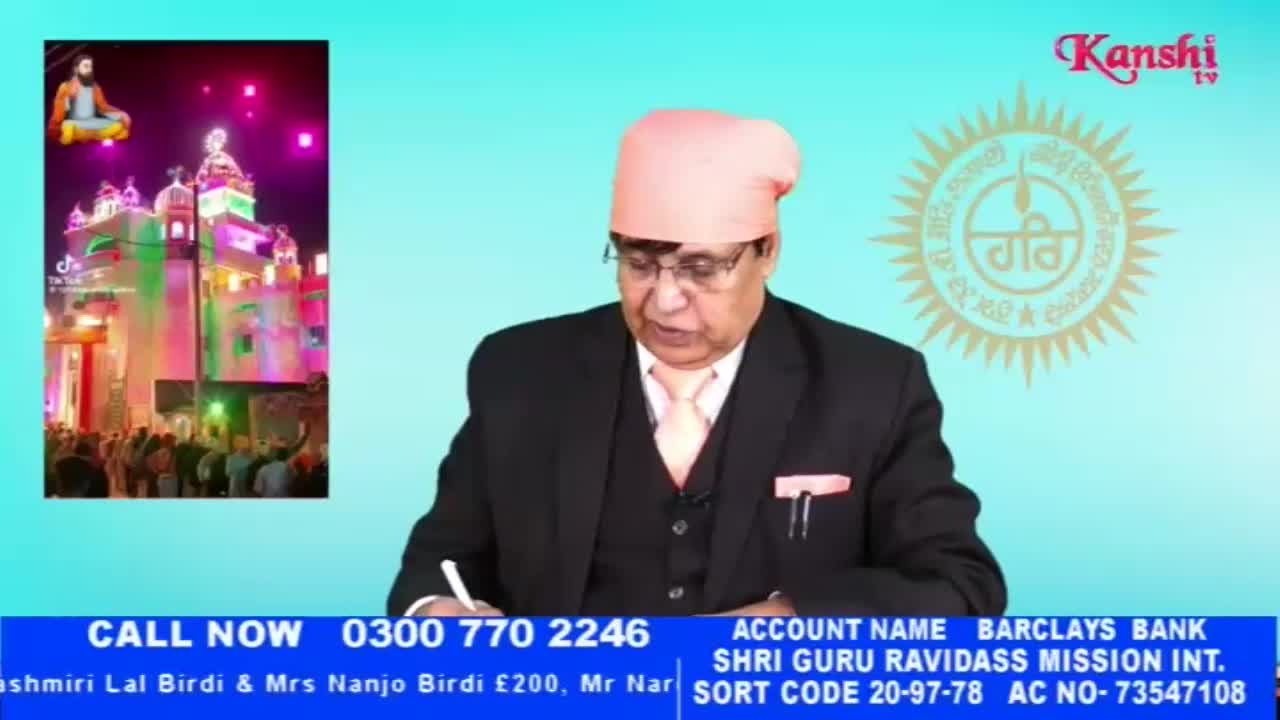 A man in a suit and turban is writing on paper. To his left, a video shows a brightly lit building with a crowd gathered outside.