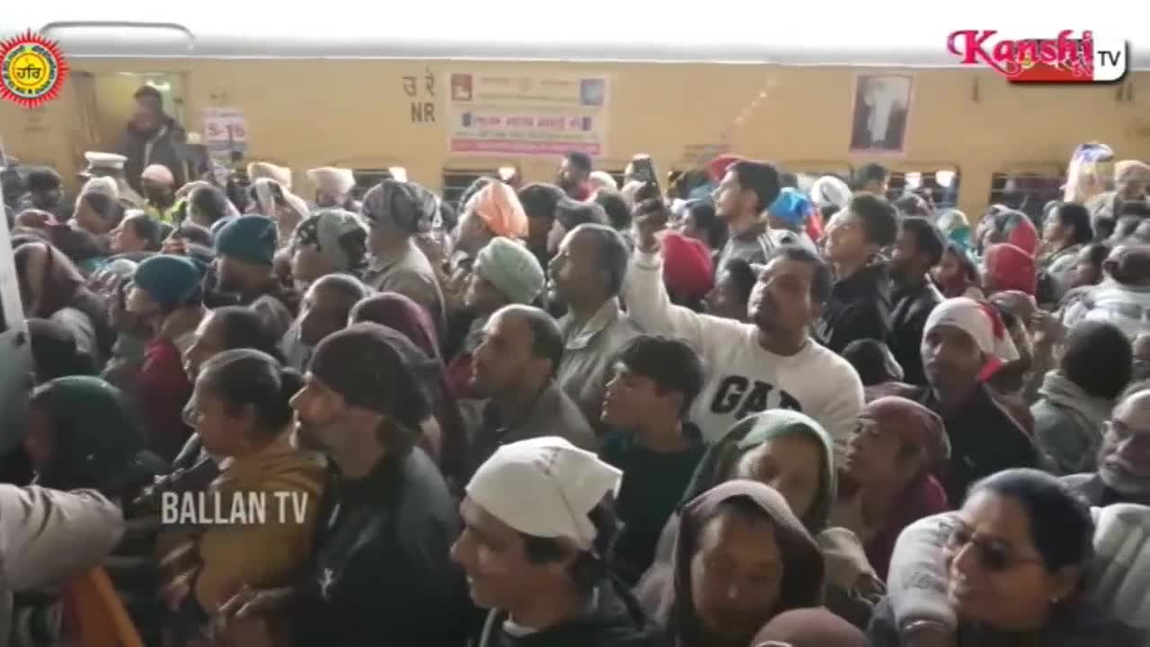 A packed train carriage waits, its doors open to a throng of people on the platform. Many are looking up, some holding phones to record, as a Kanshi TV logo appears in the corner.