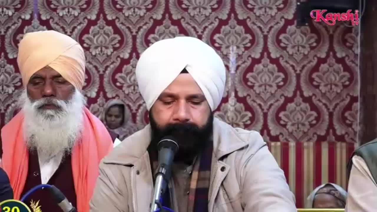 A man with a white turban and beard speaks into a microphone, his eyes closed. To his left, another man in a peach turban and orange scarf looks down. The Kanshi TV logo appears in the upper right corner.
