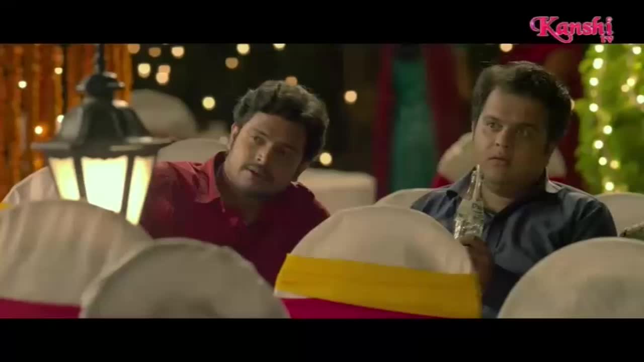 Two men sit at a table, one in a red shirt looks to his left, while the other in a blue shirt holds a bottle. The Kanshi TV logo appears in the top right corner, suggesting this is a scene from a show.