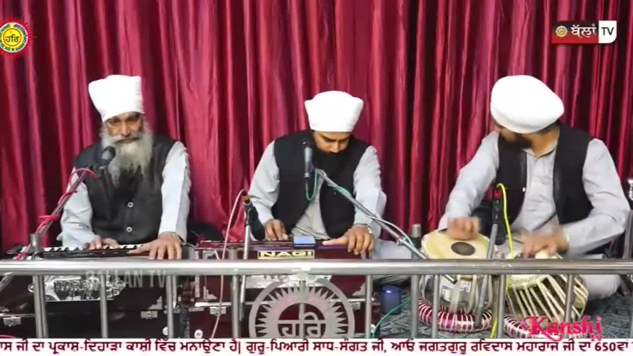 A man with a long grey beard plays a harmonium, his fingers moving across the keys. To his right, another man plays a tabla, his hands striking the drums with practiced rhythm. A third man, also wearing a white turban, plays a harmonium to the left of the tabla player.