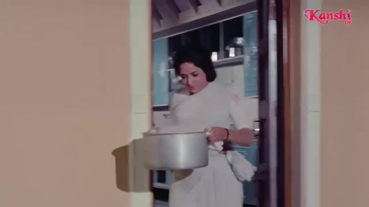 A woman in a white sari carries a large, steaming pot through a doorway. The Kanshi TV logo appears in the top right corner.