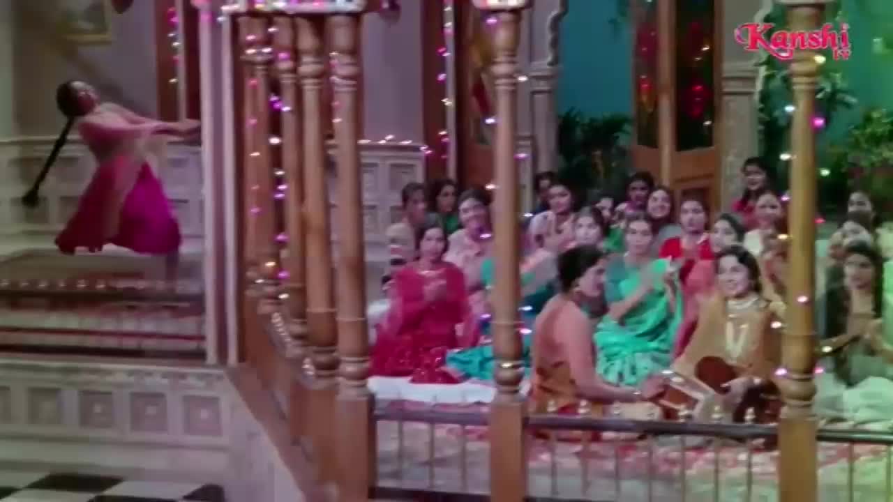 A woman in a bright pink sari is mid-leap on the stairs, her braid flying behind her. Below, a crowd of women in colourful saris watches, some clapping to the music.