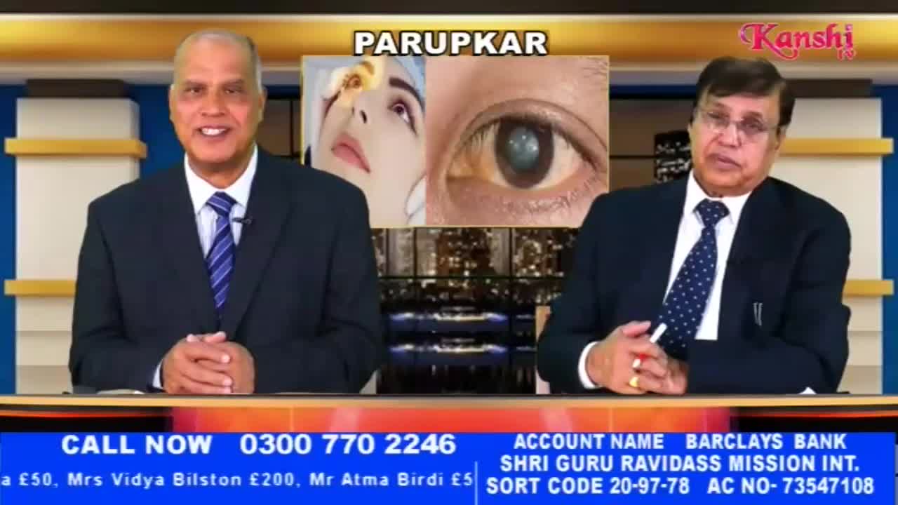 Two men in suits sit at a desk, with a graphic behind them showing a before-and-after of an eye. A phone number and bank details for the Shri Guru Ravidass Mission appear at the bottom of the screen.
