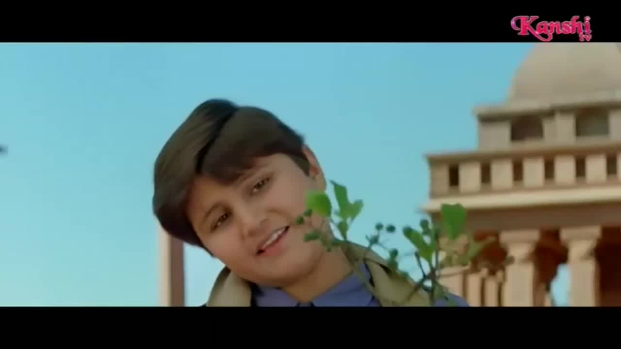 A young boy with dark hair looks to his left, a small plant obscuring part of his face. Behind him, a building with pillars and a domed roof stands against a bright blue sky.