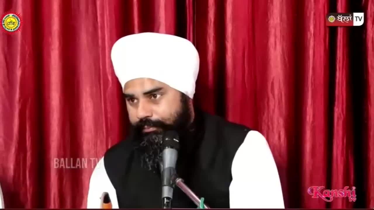 A man in a white turban and black vest speaks into a microphone. Red curtains hang behind him, with a Kanshi TV logo in the corner.