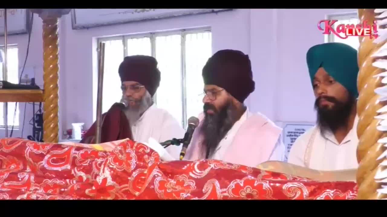 Three men in turbans sit behind a table covered in a red and white patterned cloth. A microphone is placed in front of the man in the center, and he appears to be speaking. The Kanshi TV logo is visible in the upper right corner.