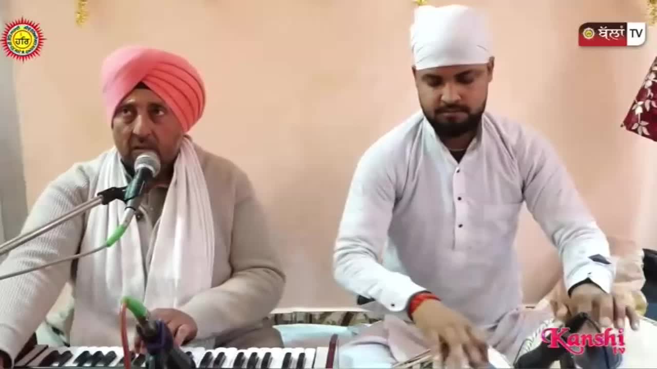 A man in a pink turban sings into a microphone while seated at a keyboard. Beside him, another man in a white head covering plays a tabla drum.