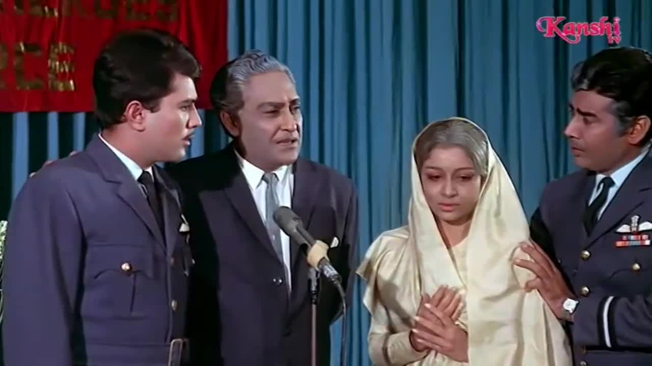 A man in a dark suit speaks into a microphone, his expression earnest. Beside him, a woman in a pale headscarf clasps her hands, her gaze downcast. Another man in a uniform, his hand resting on her shoulder, looks on with concern.