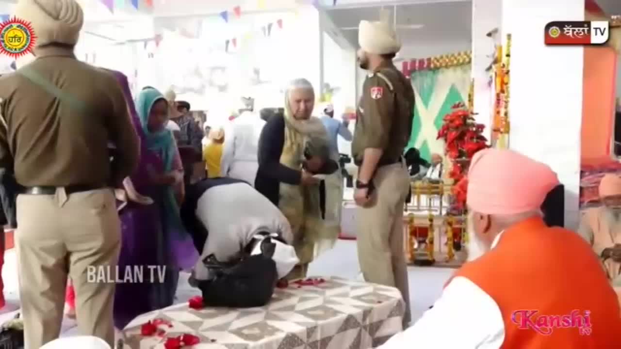A man in a grey shirt and black trousers bends low, offering something from a black bag. Two men in police uniforms stand nearby, observing the proceedings.