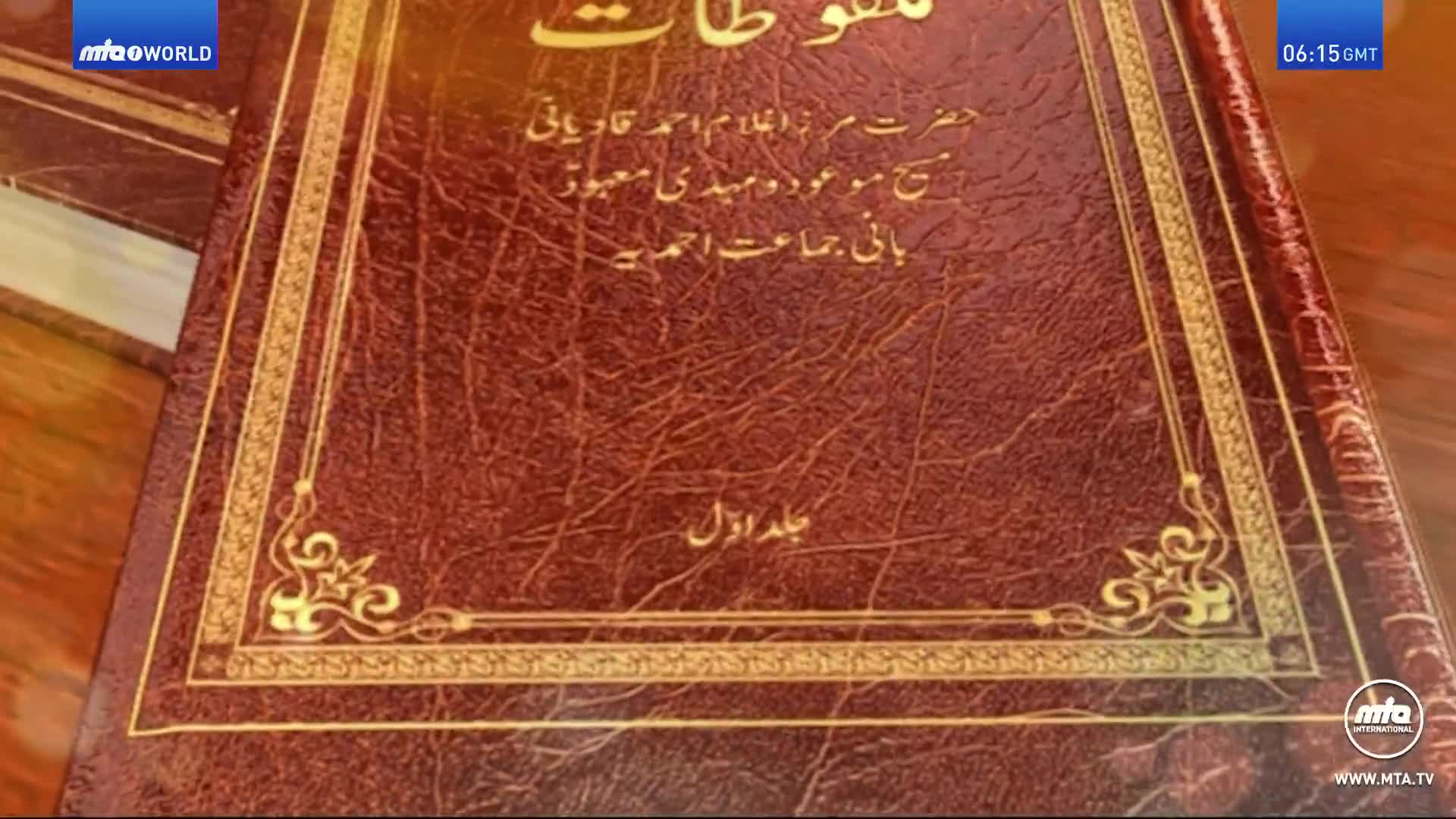 A close-up shows the cover of a book with gold lettering and ornate trim, likely part of a broadcast from MTA International in the United Kingdom. The cover is a deep red, its texture appearing like aged leather.
