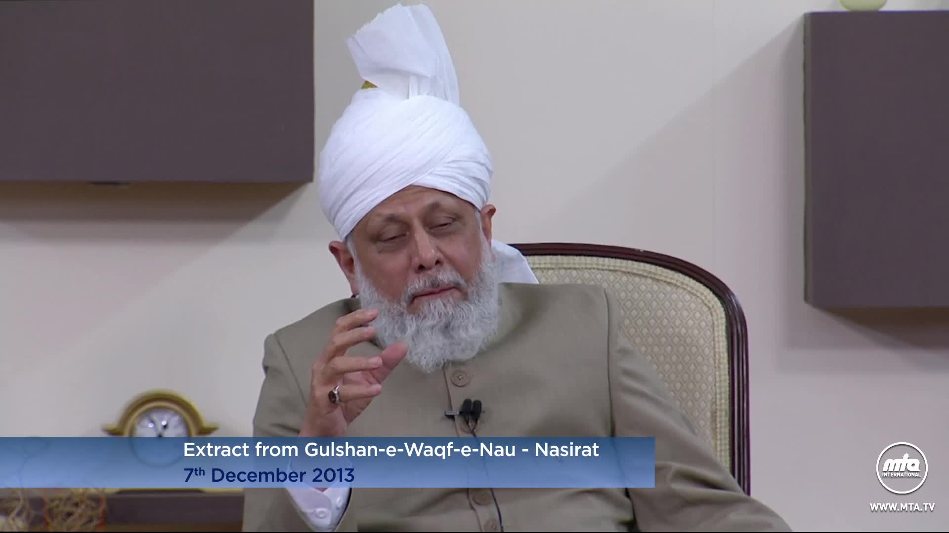 He's seated in a chair, gesturing with his right hand as he speaks. The man wears a white turban and a light-colored suit jacket.
