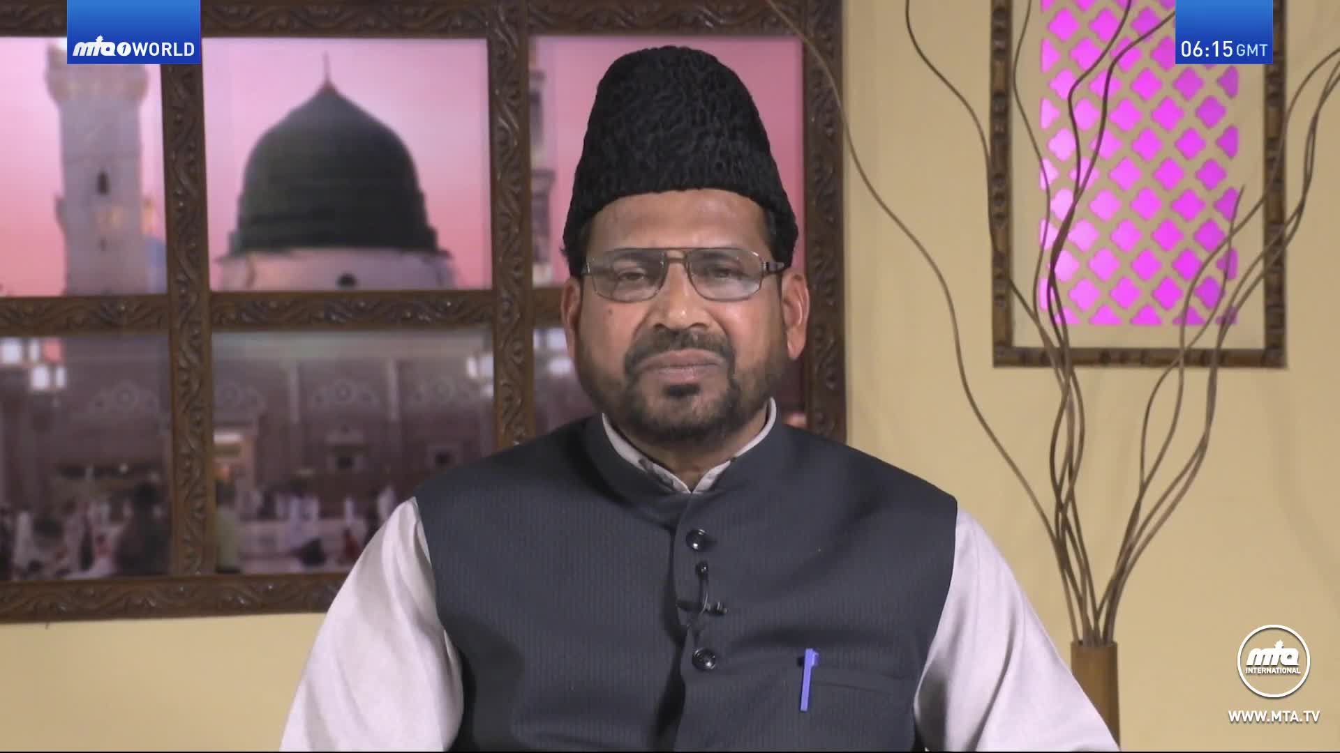 A man in a black cap and glasses is speaking directly to the camera. Behind him, a framed image of a mosque is visible on the MTA1 World channel.

