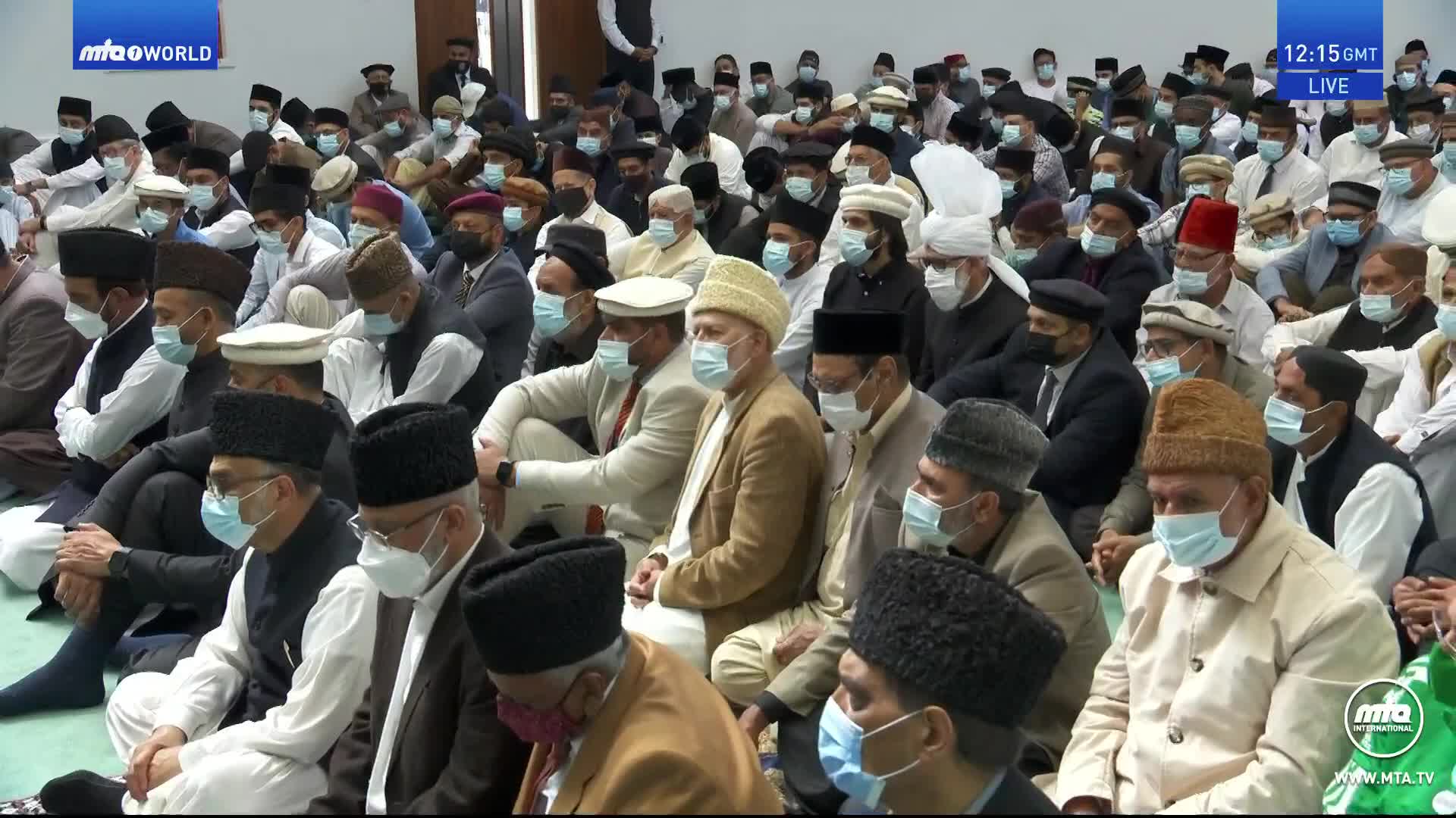 A large gathering of men, many wearing face masks and traditional headwear, sit close together on the floor. The scene is being broadcast live on MTA1 World from the United Kingdom.
