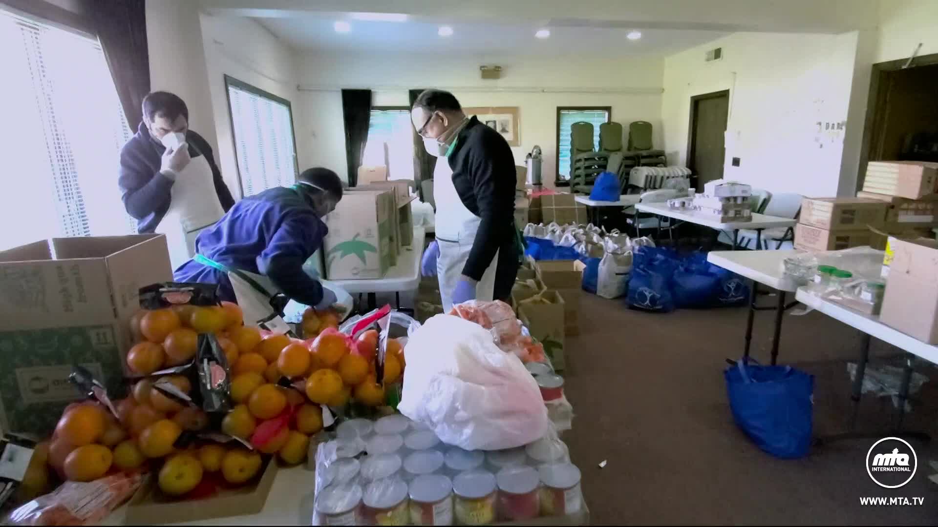 Volunteers, masked and gloved, sort oranges and other groceries onto tables.  Boxes of food and bags of supplies are stacked around the room.
