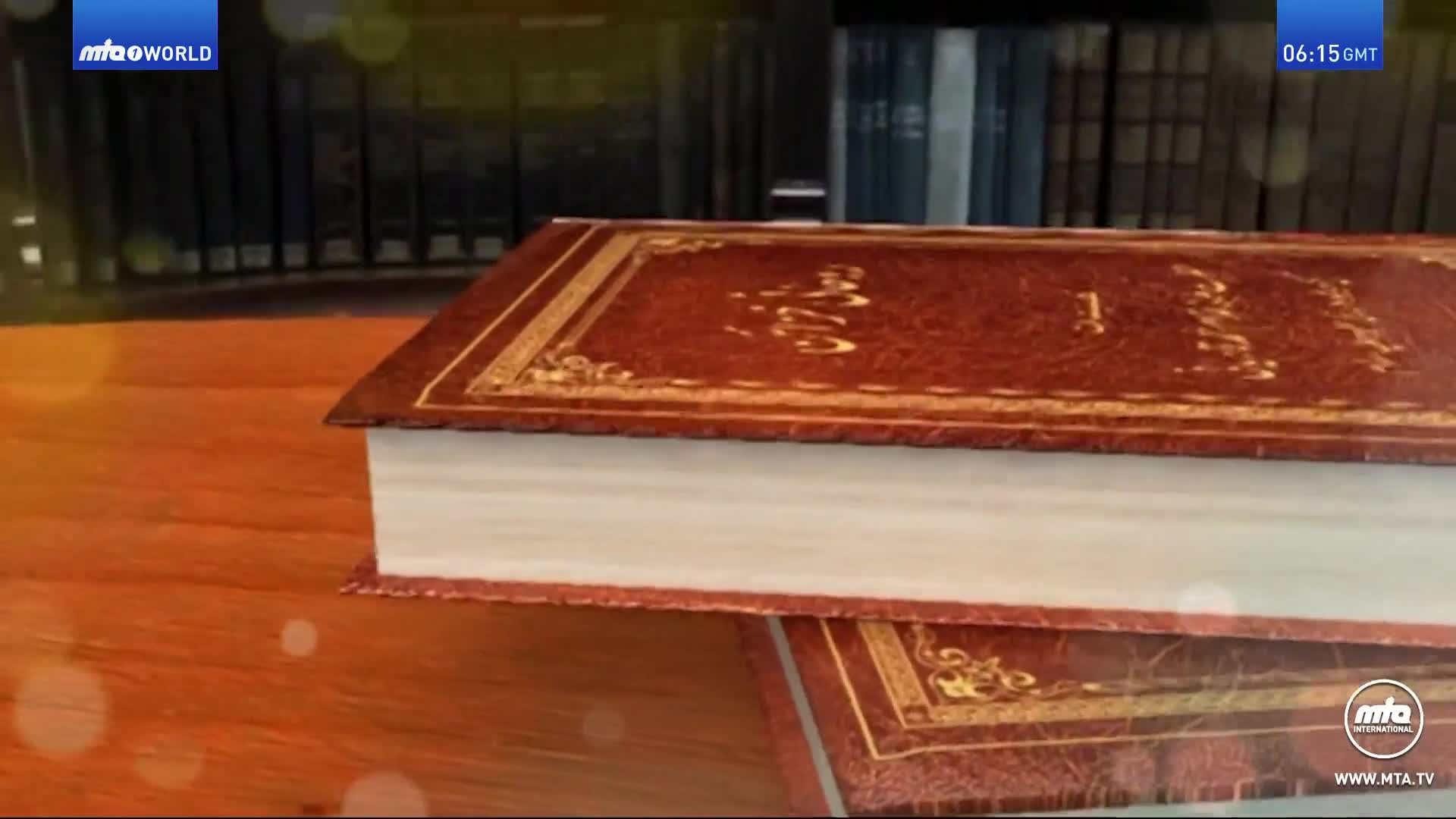 A large, ornate book rests atop another, both with brown leather covers and gold detailing. The MTA World logo and the time, 06:15 GMT, are visible in the corner.
