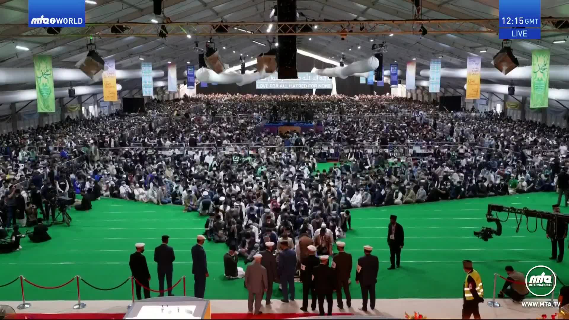 Thousands of people are gathered on the green carpet, many wearing white hats, all facing the front. A group of men in dark suits and white hats stand on a raised platform, looking out at the crowd.
