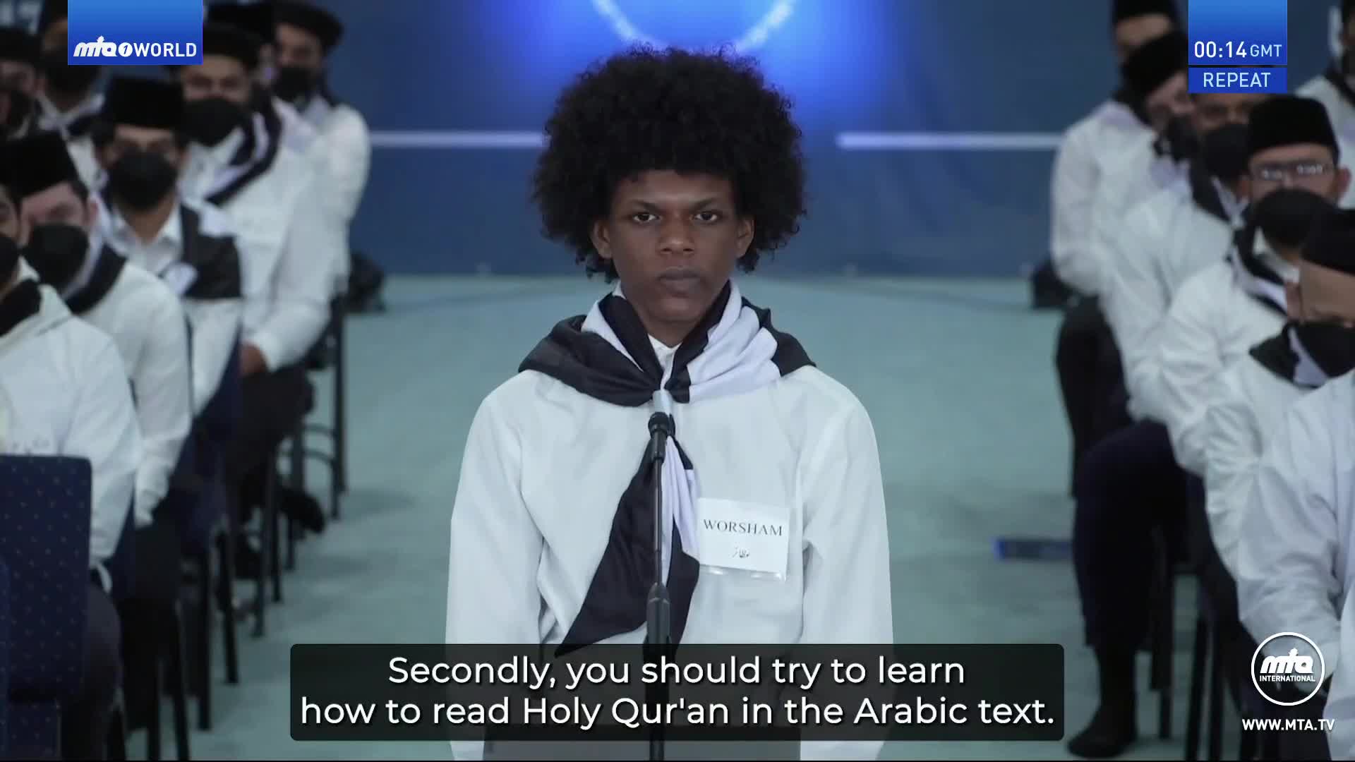 A young man with a large afro stands at a microphone, a black and white scarf draped over his shoulders. Behind him, a group of people in white shirts and black hats sit in rows.
