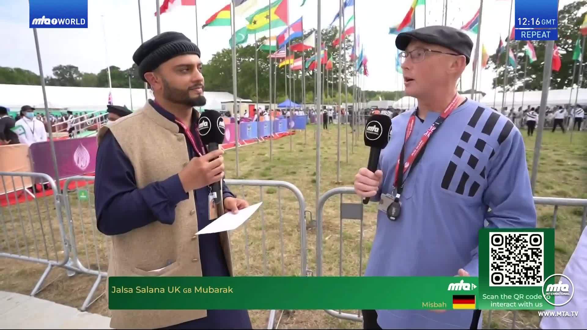 Two men, each holding a microphone, are speaking at the Jalsa Salana UK event. Behind them, rows of flags wave in the breeze, and a crowd of people are gathered in the distance.
