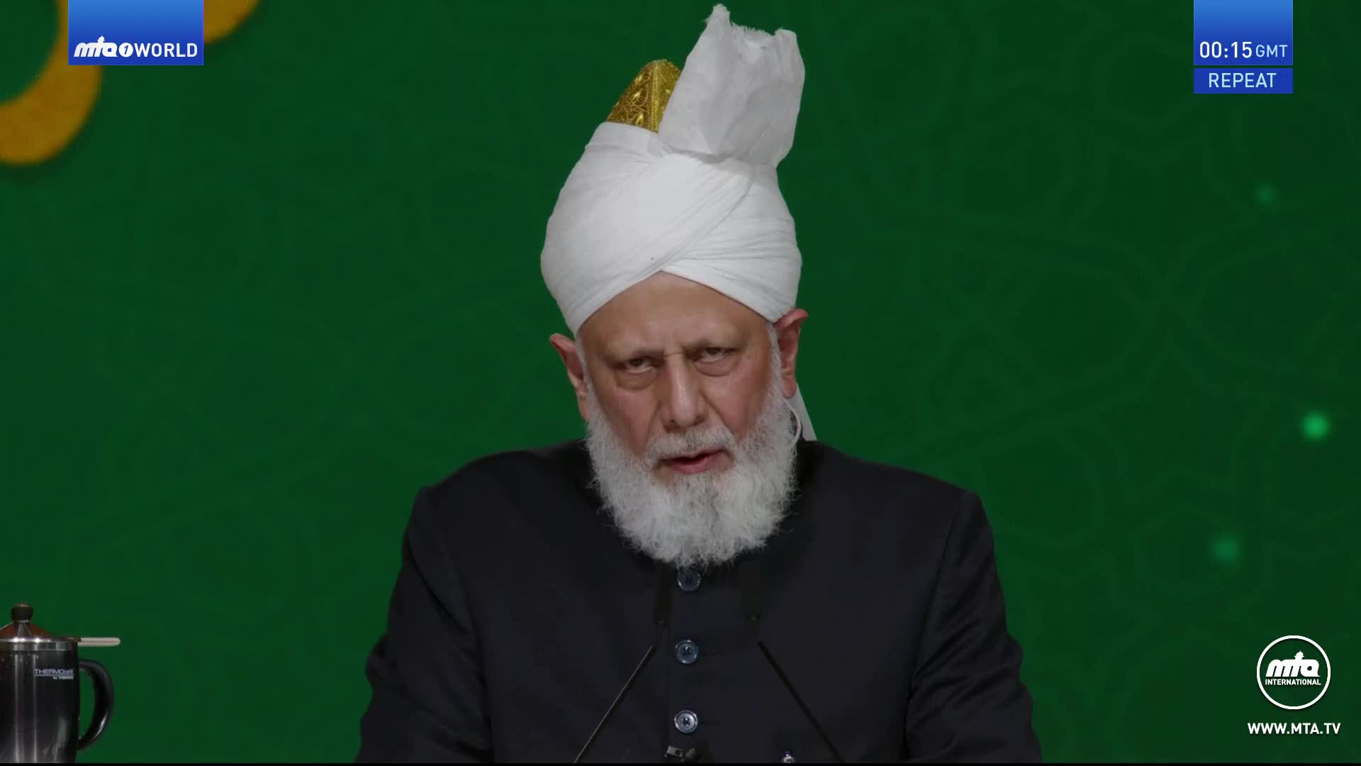 The man in the white turban is speaking, his gaze direct. Behind him, a green backdrop and the MTA1 World logo are visible.
