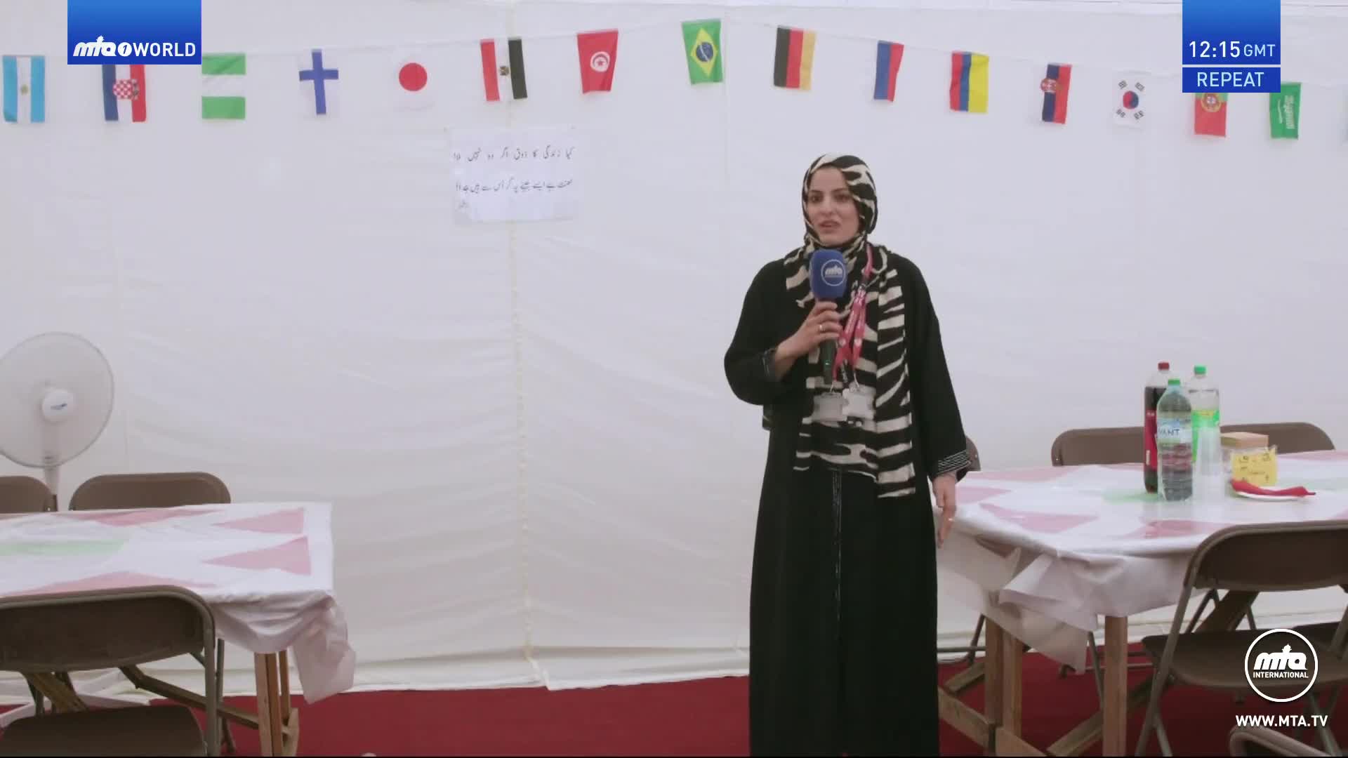 A woman in a black dress and zebra-striped scarf holds a microphone, speaking in front of a white backdrop decorated with flags. Tables covered with white tablecloths are set up, and a fan sits nearby.
