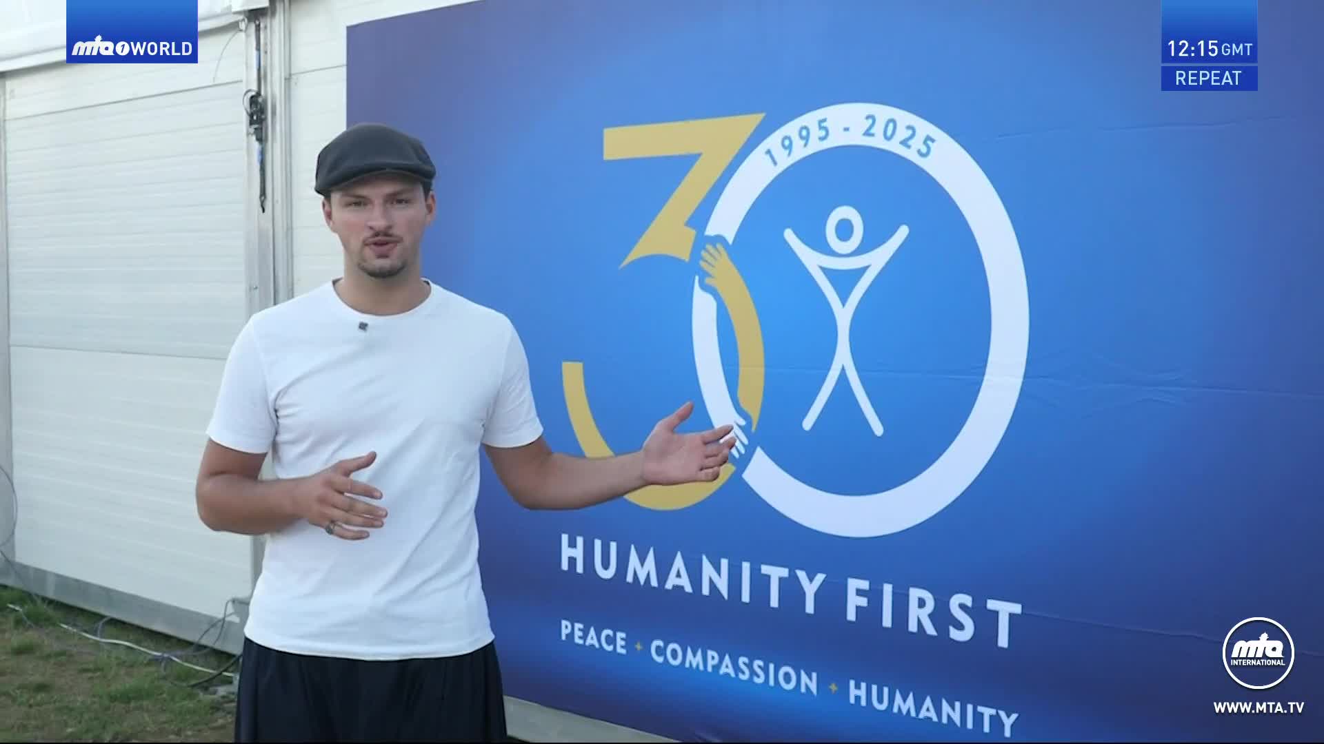 A man in a white t-shirt and cap gestures towards a large blue banner with the MTA International logo and the words "Humanity First." He's standing outside, likely in the United Kingdom, with the MTA1 World logo visible in the corner.
